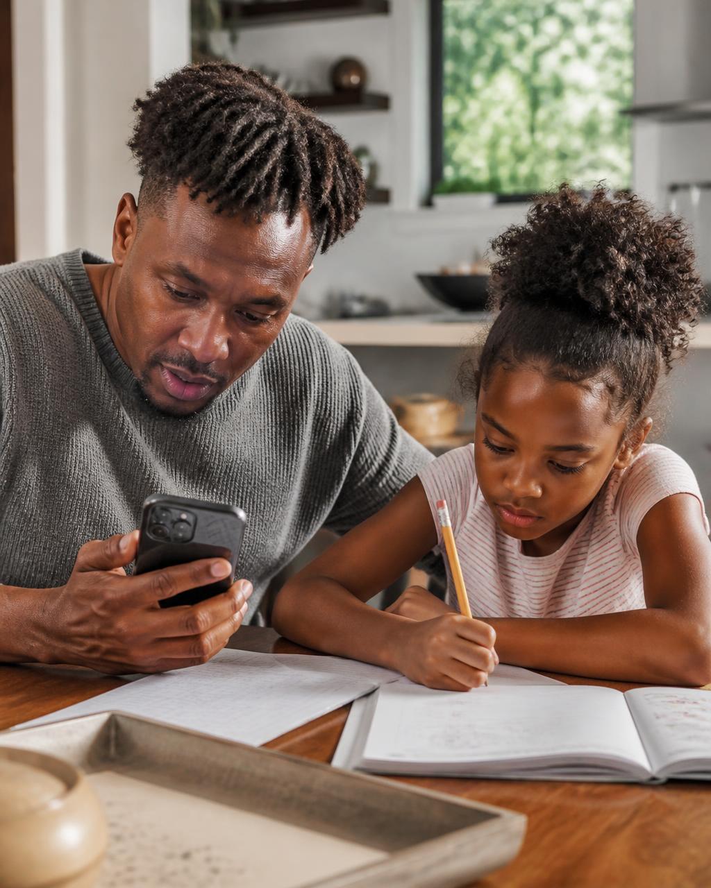 A father sitting beside his daughter during homework, looking something up on his phone